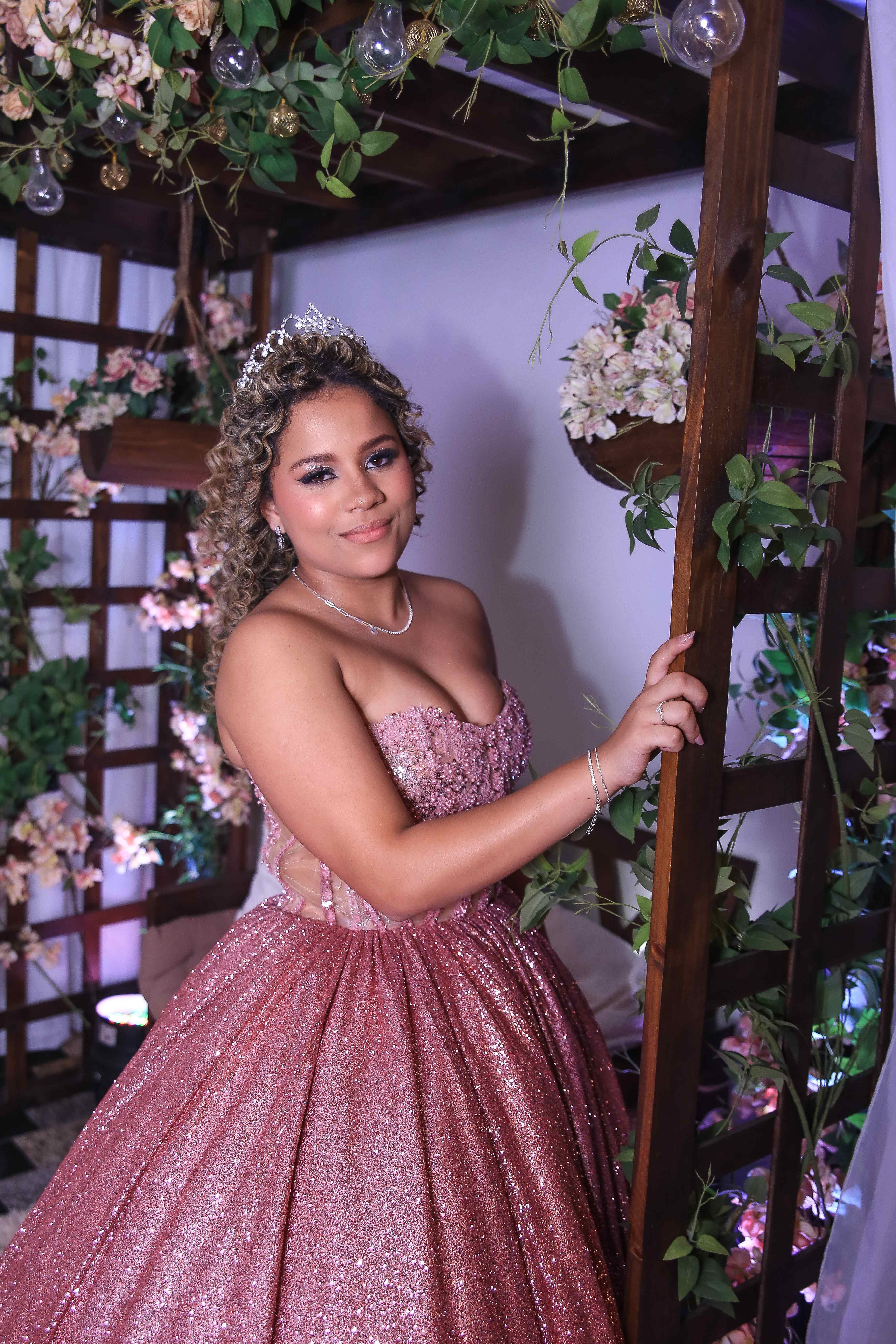 Debutante de cabelo cacheado com tiara e vestido rosa cintilante, ao lado de treliça com flores
